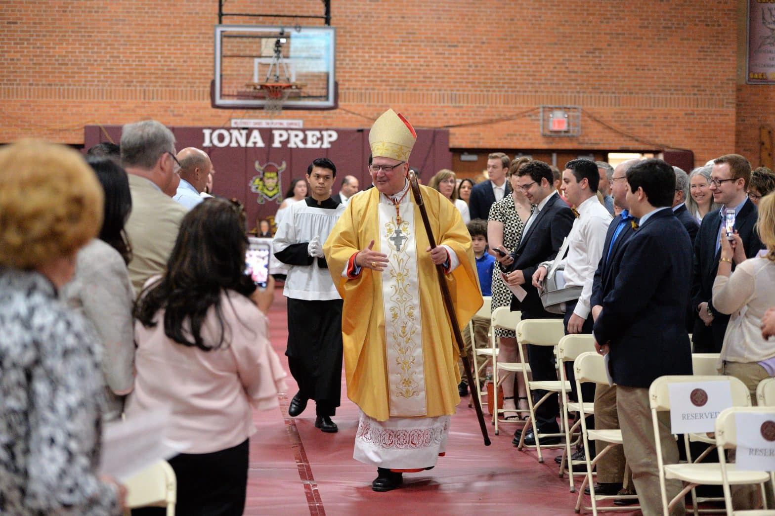 His Eminence, Timothy Cardinal Dolan, followed by altar server and Iona Prep 11th grader Ian Mendoza of Cortlandt Manor, greets Iona Preparatory students, alumni, families and friends during a special Centennial Mass in the Tully Gymnasium